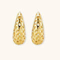 Close-up of small gold-plated hoop earrings with intricate honeycomb cutout pattern on cream background.